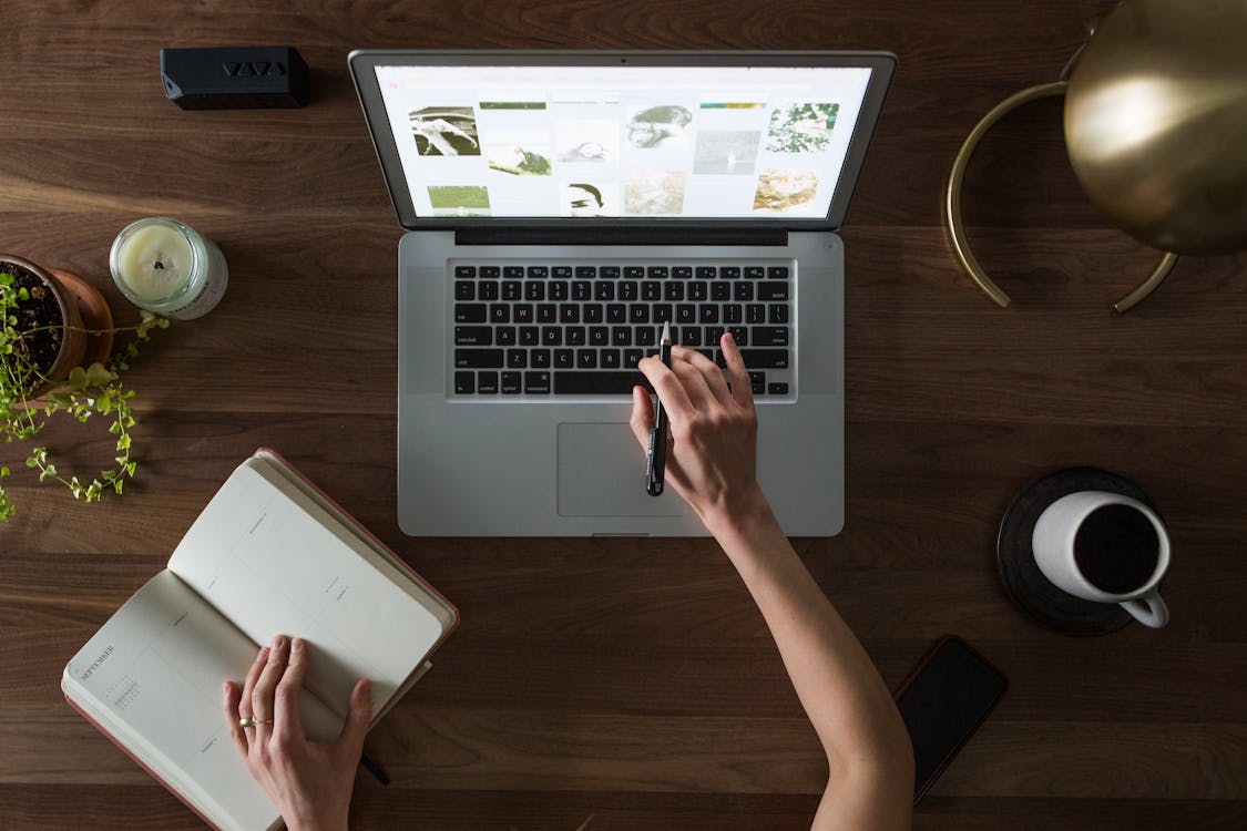 An Image of a Person Using Macbook While Holding on Planner Book