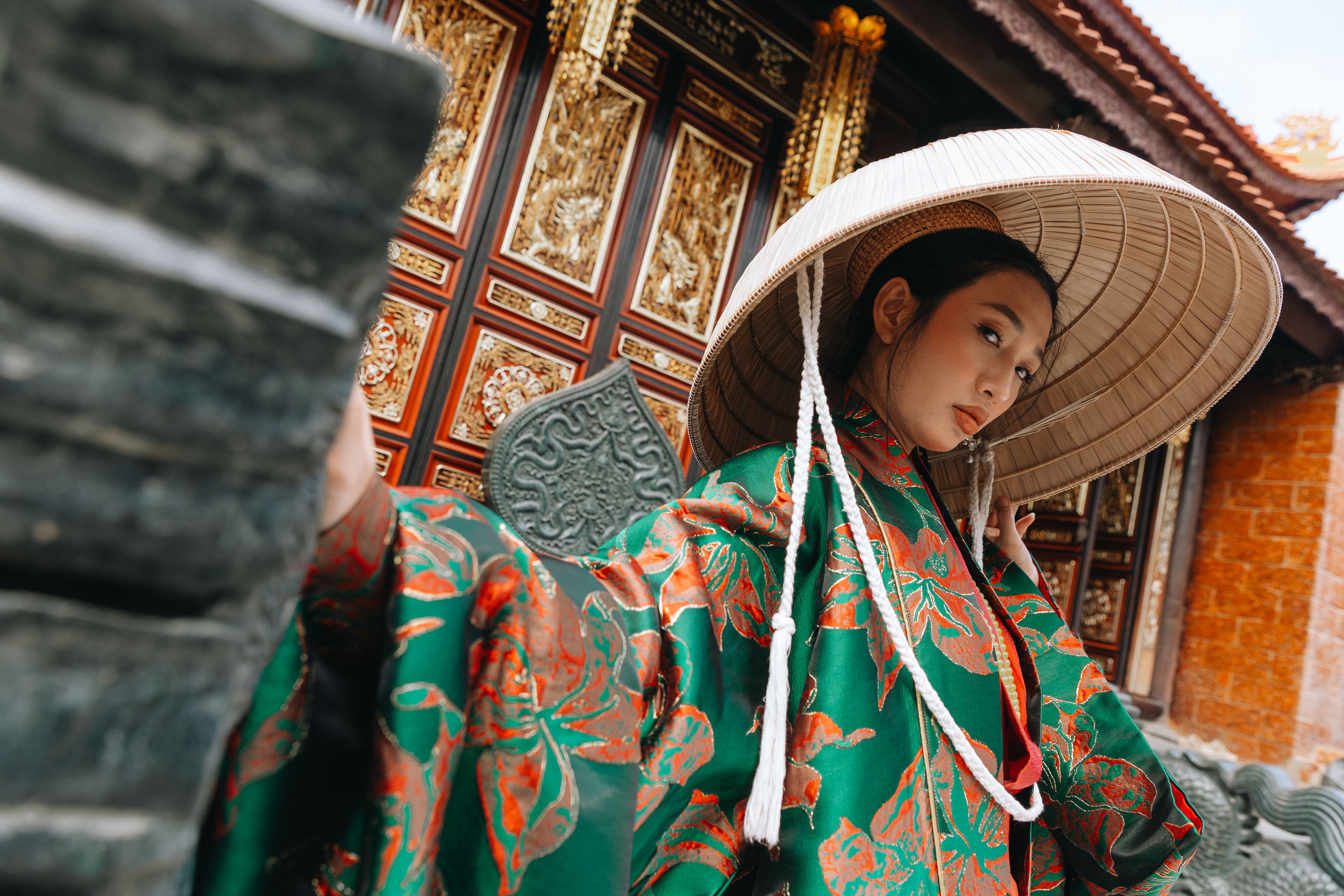 Portrait of a woman in traditional Vietnamese dress and conical hat in front of ornate temple doors in Hanoi.