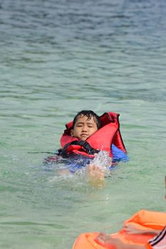 Young boy in a red life jacket swimming in clear waters, enjoying a sunny summer day.