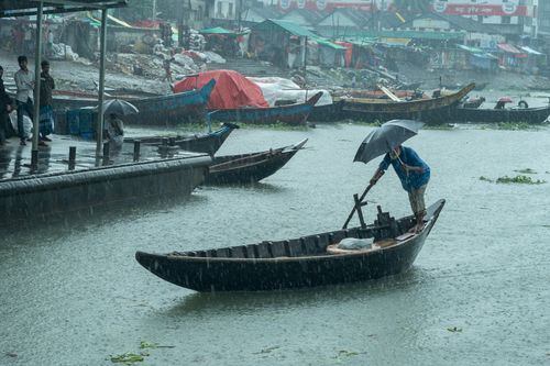 Free A man navigates a boat in the rain with an umbrella at a bustling river port. Stock Photo