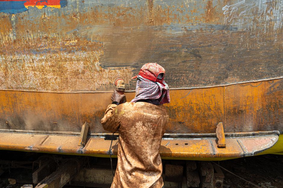 A worker sands a rusty metal surface outdoors using a power tool, wearing protective clothing.
