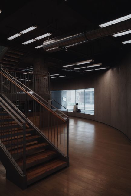 Dimly lit modern interior with a solitary figure by the window, featuring industrial stairs.