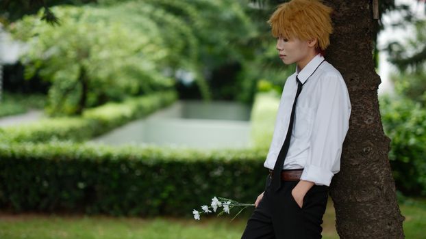 A young person in formal attire leans against a tree holding flowers, in a serene garden setting.
