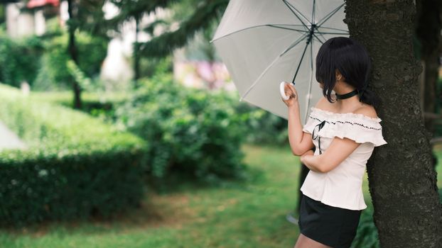 A woman standing against a tree with an umbrella in a green garden setting, exuding a serene and peaceful mood.
