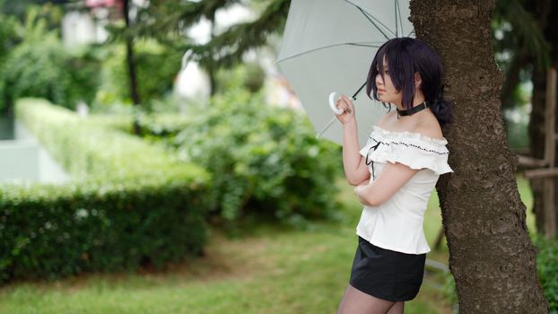 Young woman in casual summer attire standing under a tree with an umbrella in a lush garden.