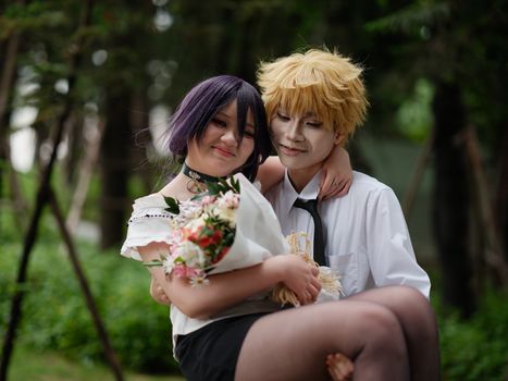 Two cosplayers share a tender moment outdoors, surrounded by greenery and holding a bouquet.