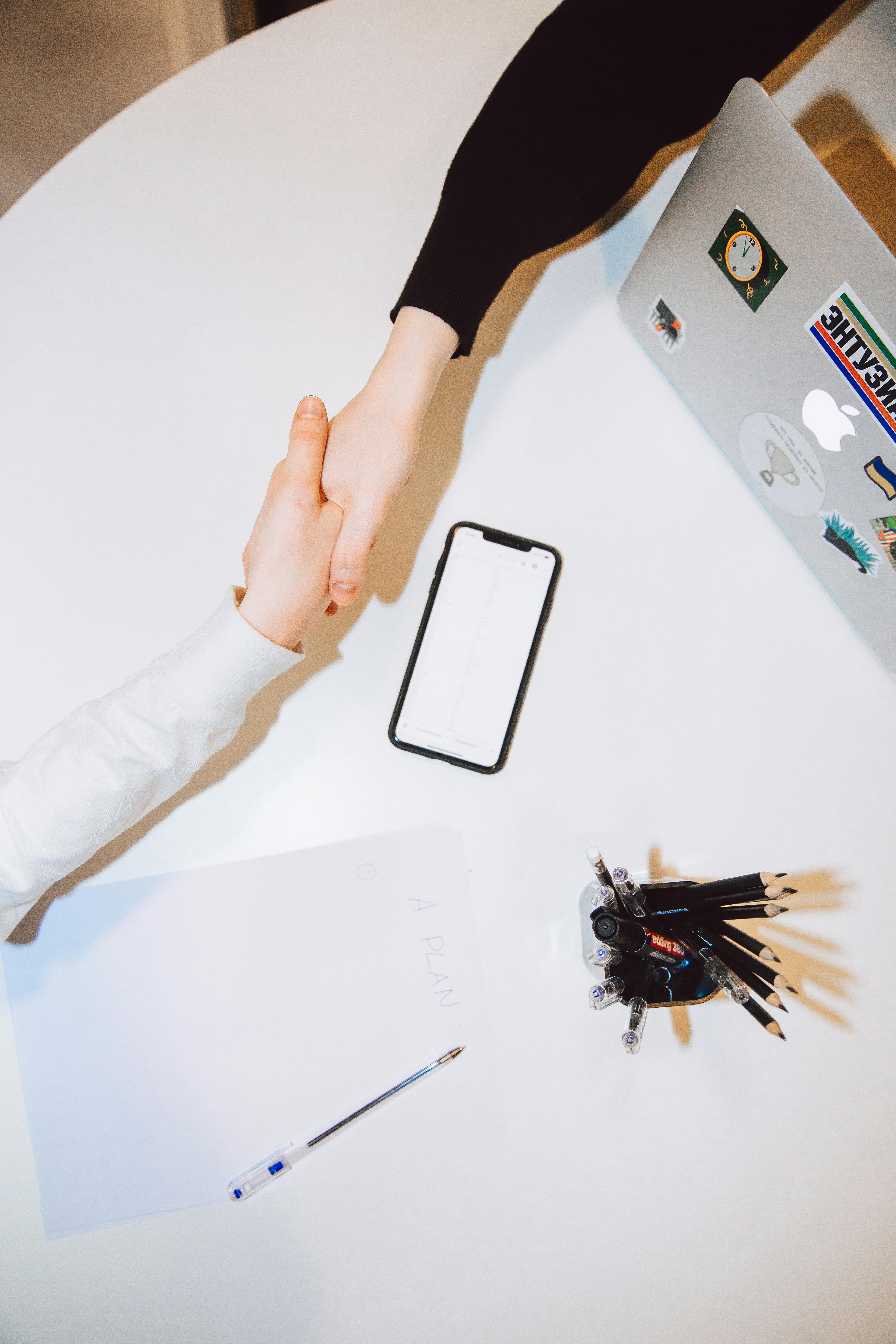 Two people shaking hands at a desk with laptop, phone and documents"