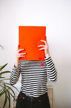 一位年轻女子穿着条纹衬衫，用一本亮橙色的书遮住了脸