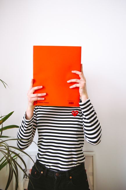 Young woman wearing a striped shirt hides face with a bright orange book.