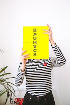 一名年轻女子穿着条纹衬衫，捂着脸，手里拿着一本大胆的黄色包豪斯书