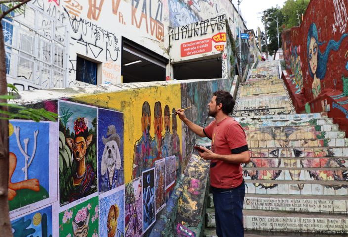 Street artist works on colorful mural in graffiti-filled urban staircase.