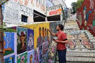 Street artist works on colorful mural in graffiti-filled urban staircase.