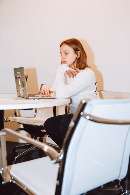 Женщина в белом топе с длинным рукавом сидит на стуле и работает на ноутбуке