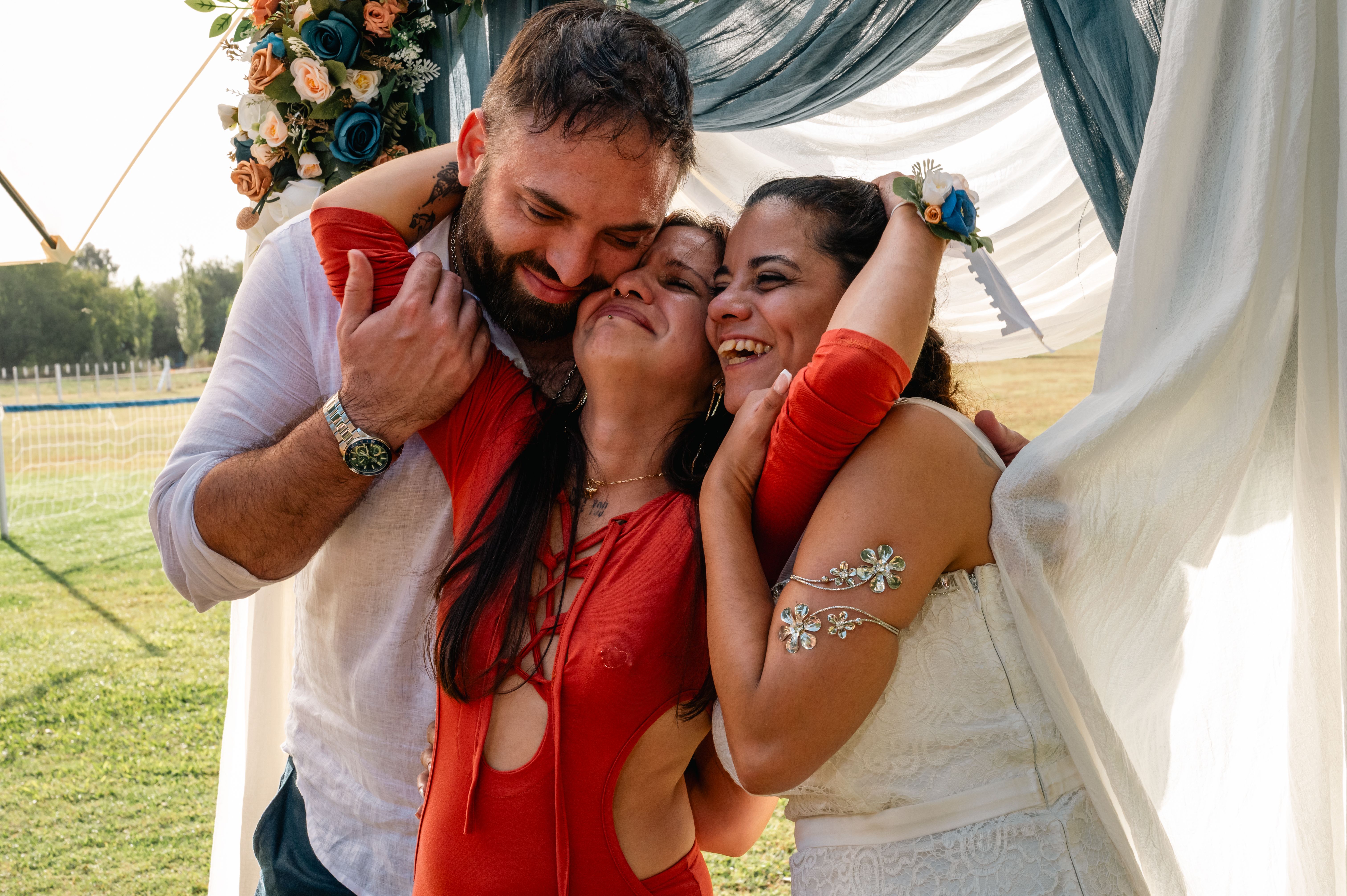 A happy trio embraces under a floral archway at an outdoor wedding celebration.