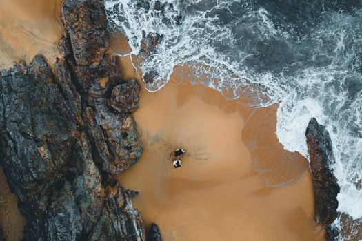 Aerial shot of a couple on a rocky beach with ocean waves crashing. Ideal for travel or lifestyle themes.