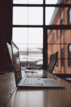 极简主义风格的办公室里，木桌上放着一台笔记本电脑，大窗户提供了充足的自然光线