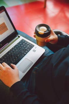 一个人拿着可重复使用的咖啡杯，在笔记本电脑上打字，展现了现代远程工作的场景