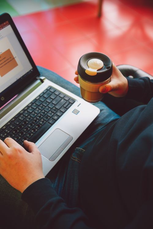 Free A person types on a laptop while holding a reusable coffee cup, illustrating modern remote work. Stock Photo