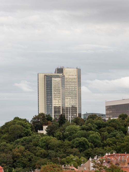 Free A tall glass skyscraper stands amidst dense greenery under a cloudy sky, showcasing urban architecture. Stock Photo