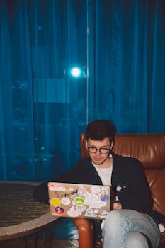 一位戴着眼镜的年轻人在室内，用一台蓝色窗帘图案的笔记本电脑工作，非常适合远程办公的主题