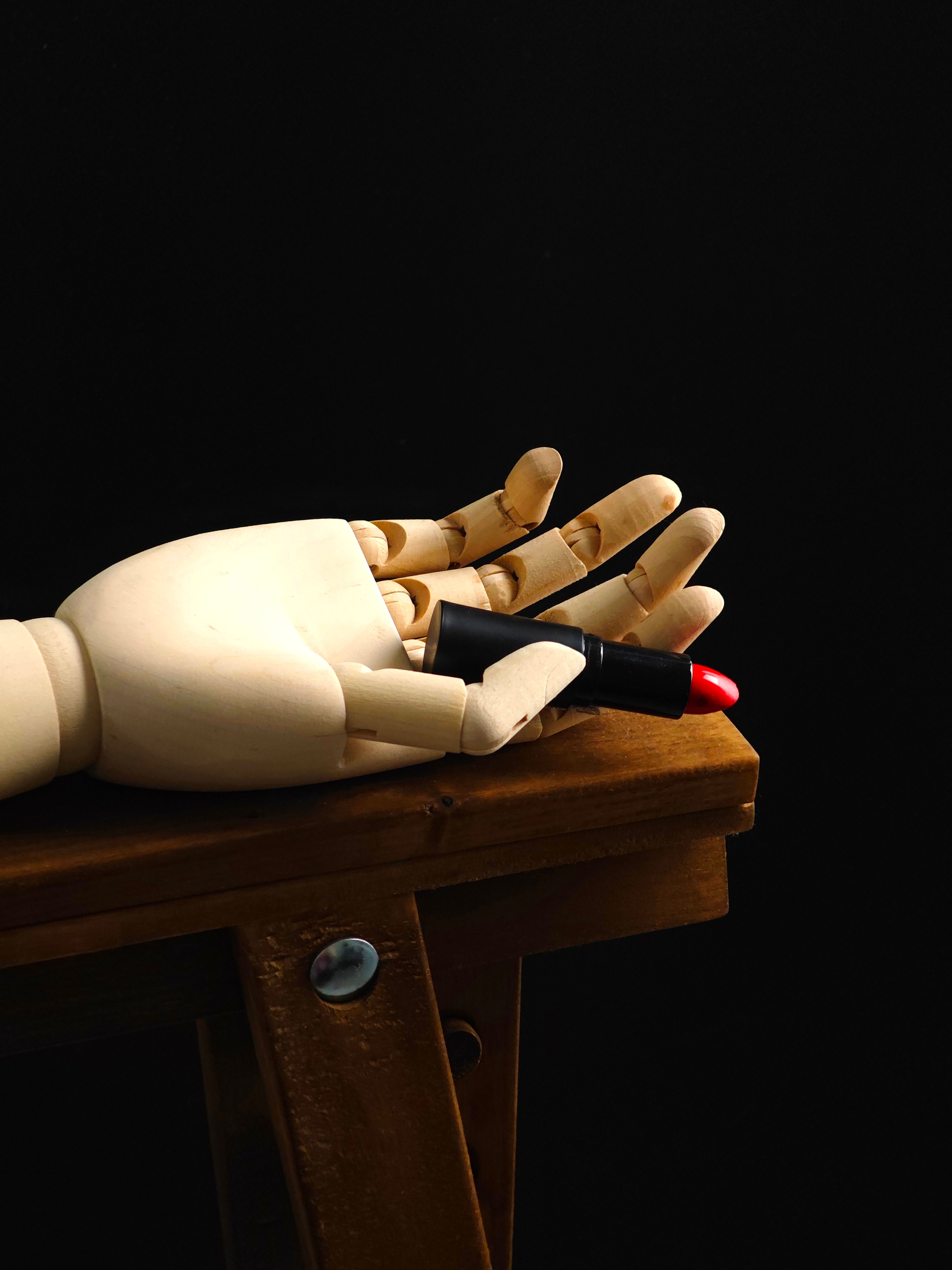 A wooden mannequin hand holding a vibrant red lipstick, placed on a rustic wooden stool against a dark background.