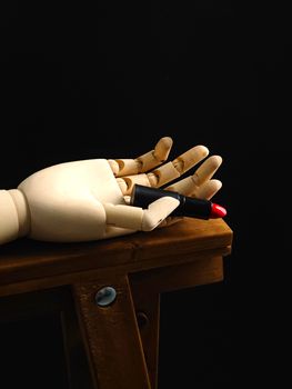 A wooden mannequin hand holding a vibrant red lipstick, placed on a rustic wooden stool against a dark background.