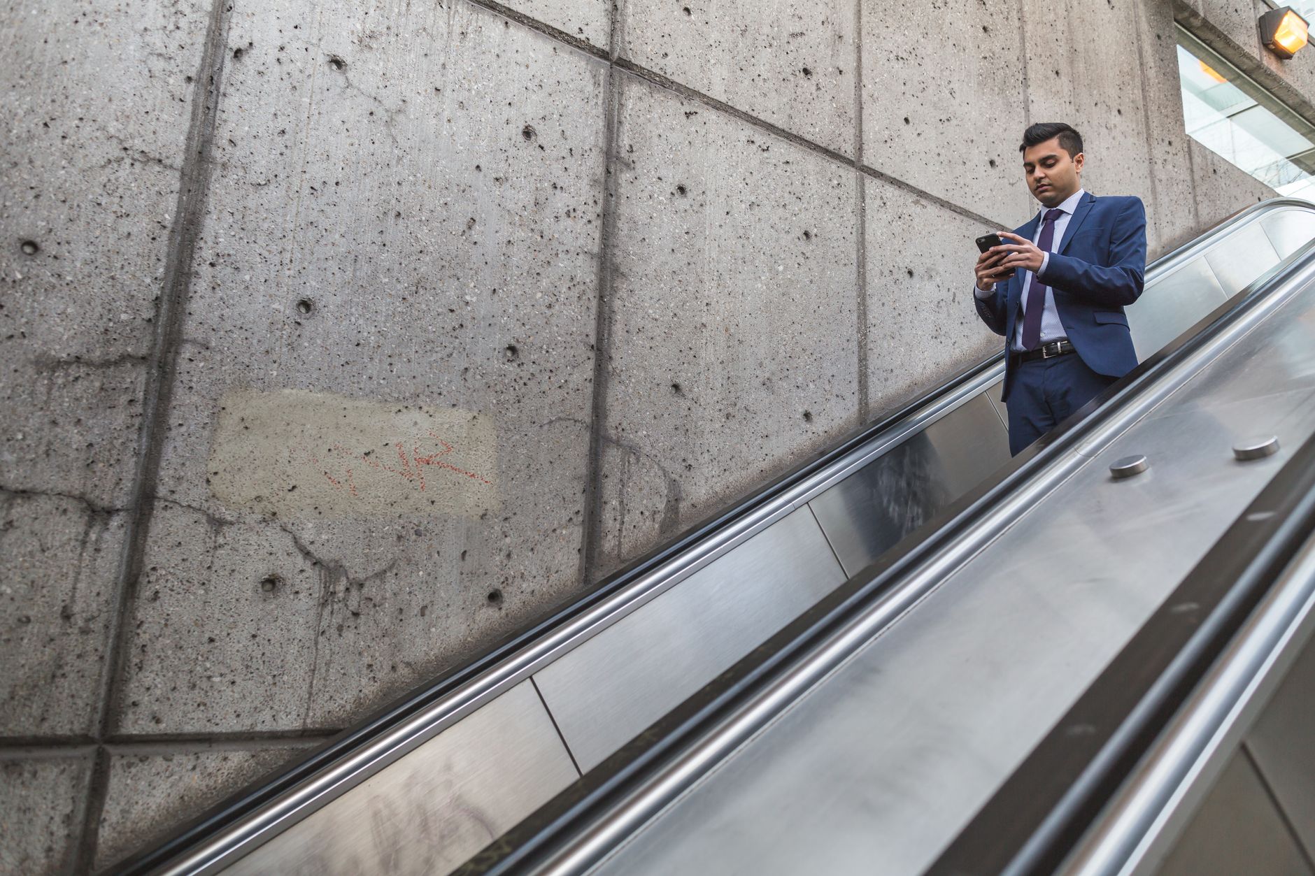 A businessman in a blue suit using a smartphone while descending an escalator indoors.