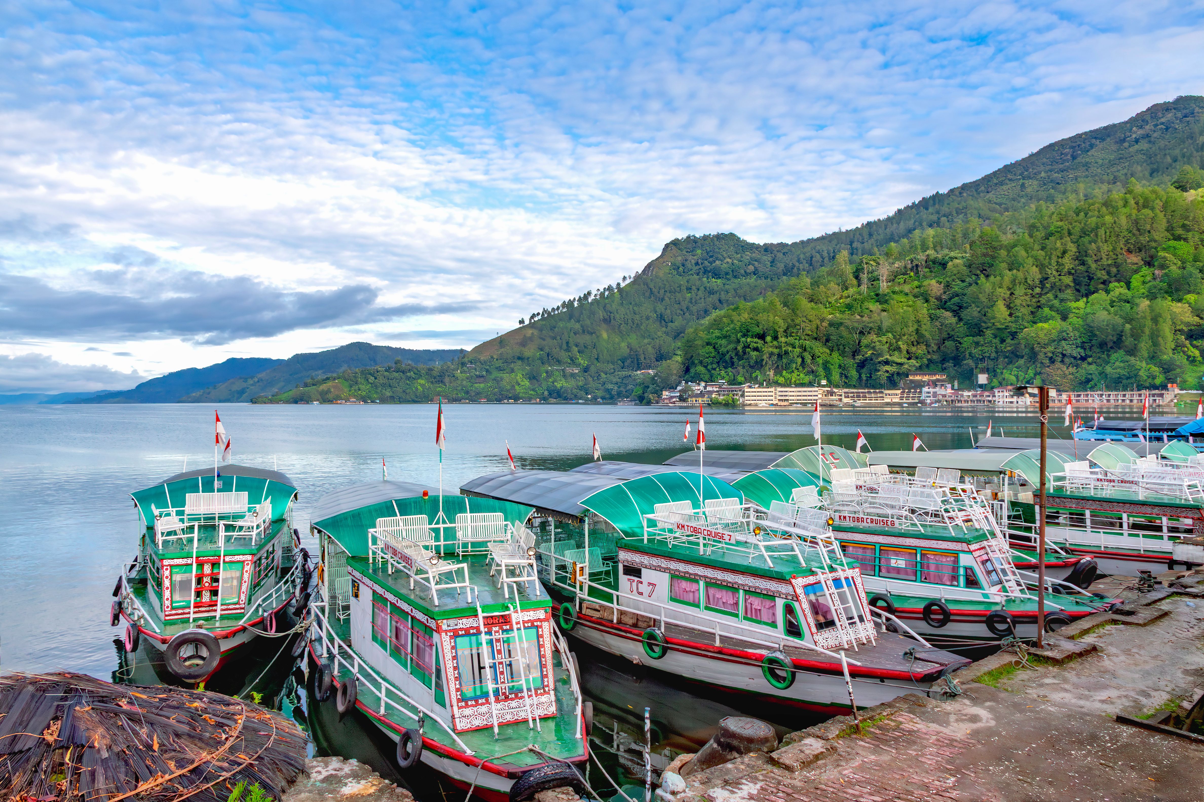 Scenic view of vibrant boats docked on Lake Toba, Indonesia, surrounded by lush hills under a bright sky.