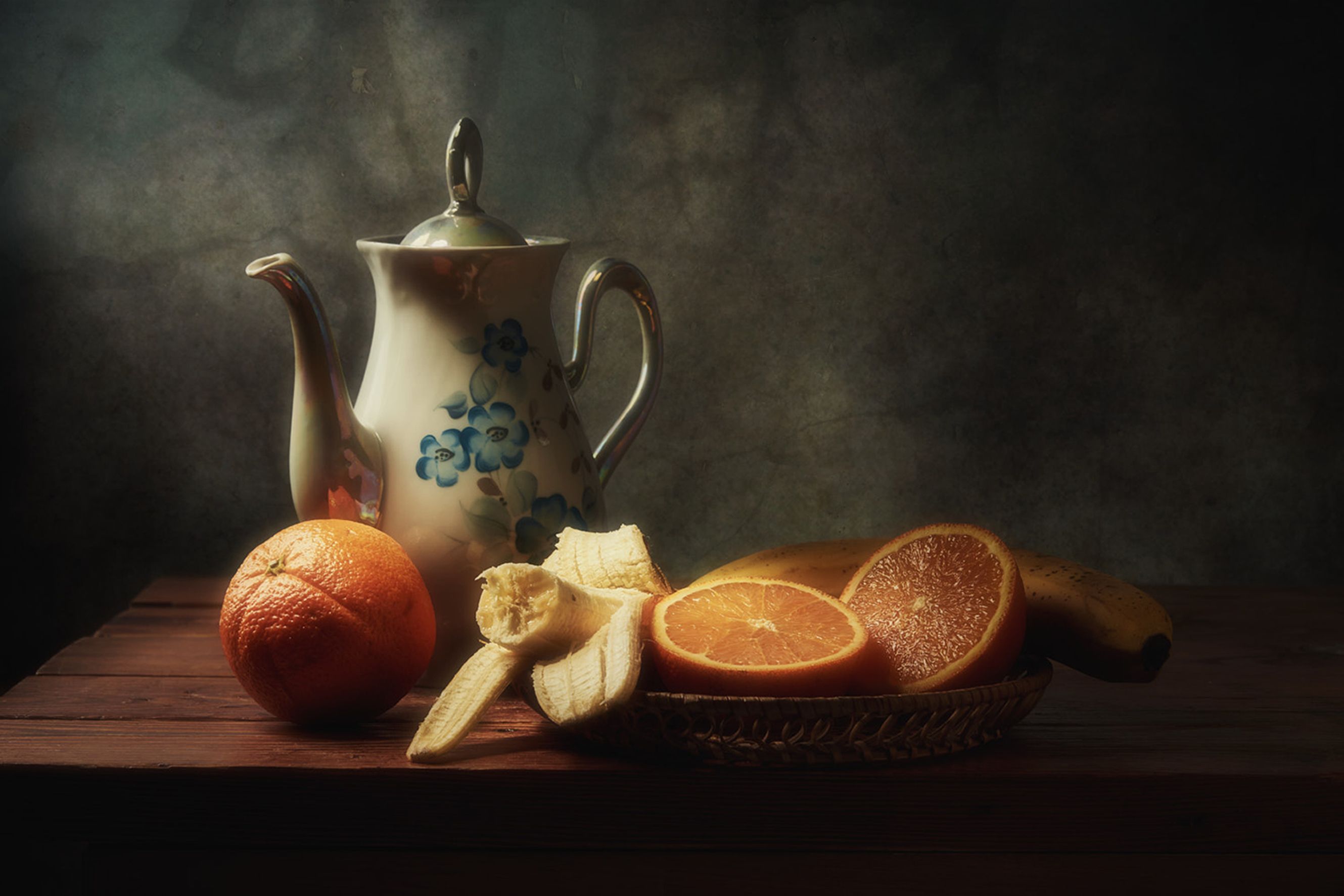 Artistic still life featuring a vintage teapot with bananas and oranges on a wooden table.
