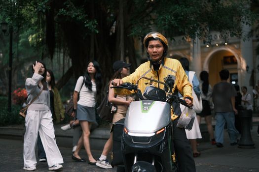 A bustling city street with a delivery rider on a scooter among pedestrians.