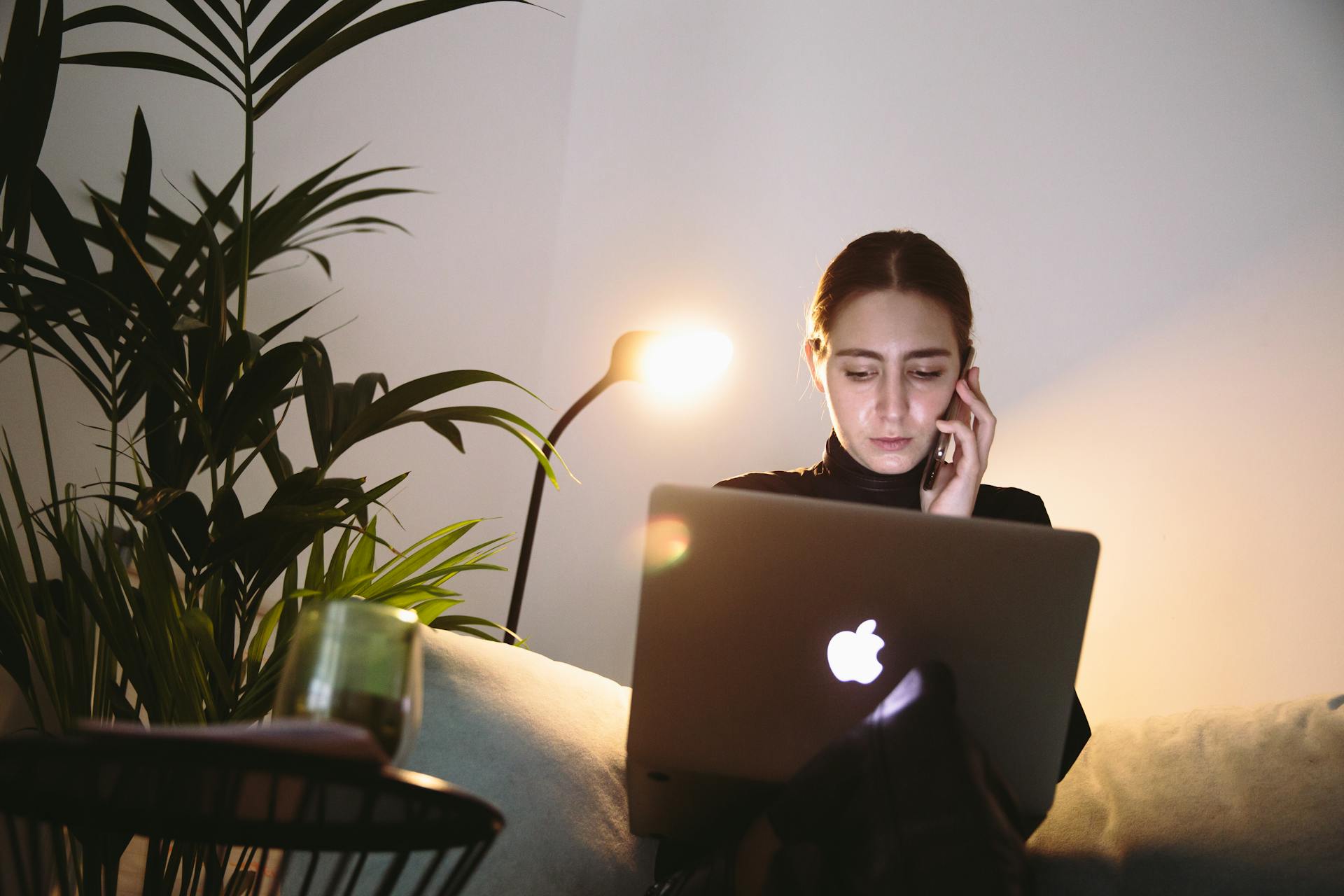 Woman in home office setting working remotely with a laptop and smartphone under lamp light.