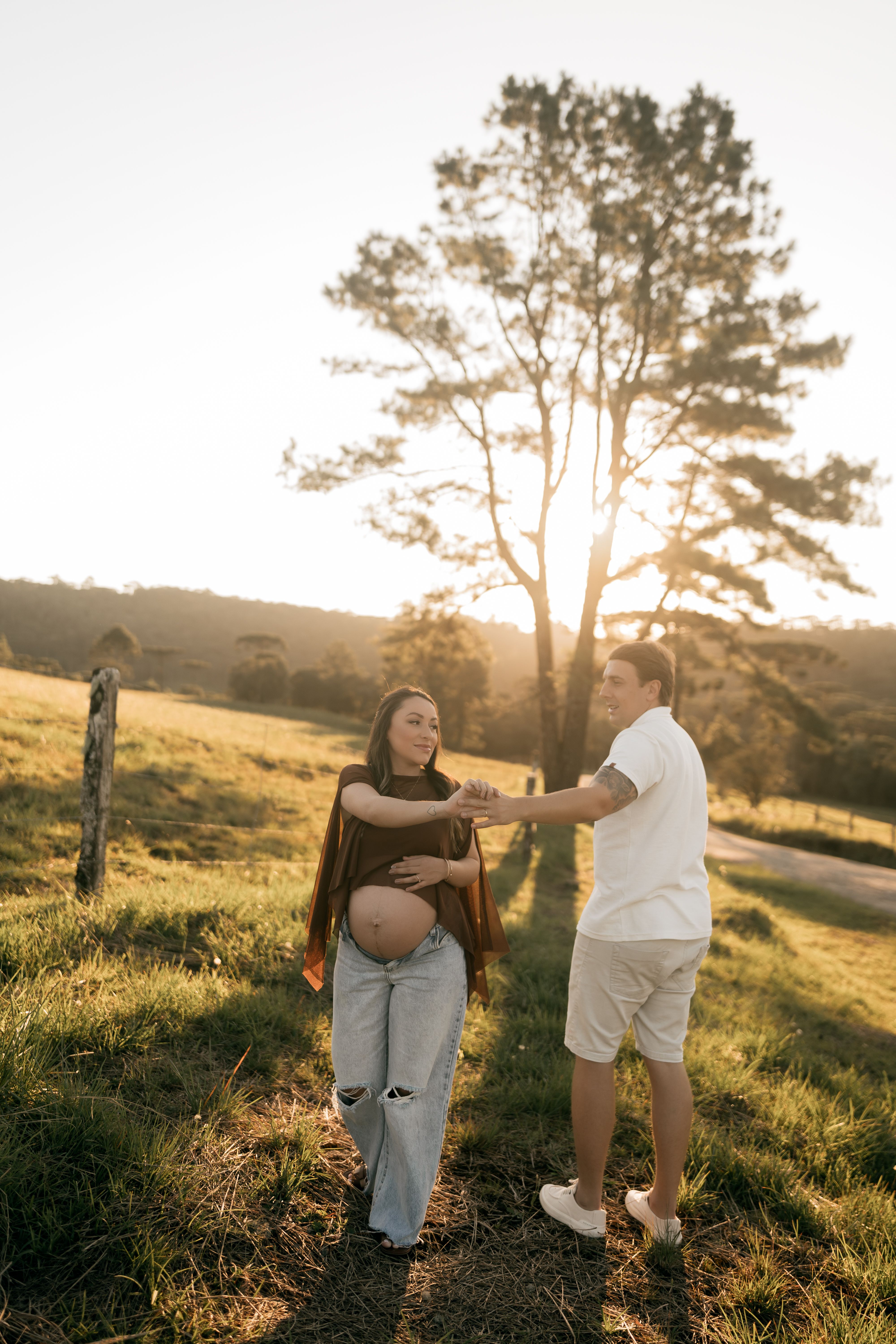 A pregnant couple dances joyfully in a scenic sunlit field, capturing a tender moment of love and anticipation.