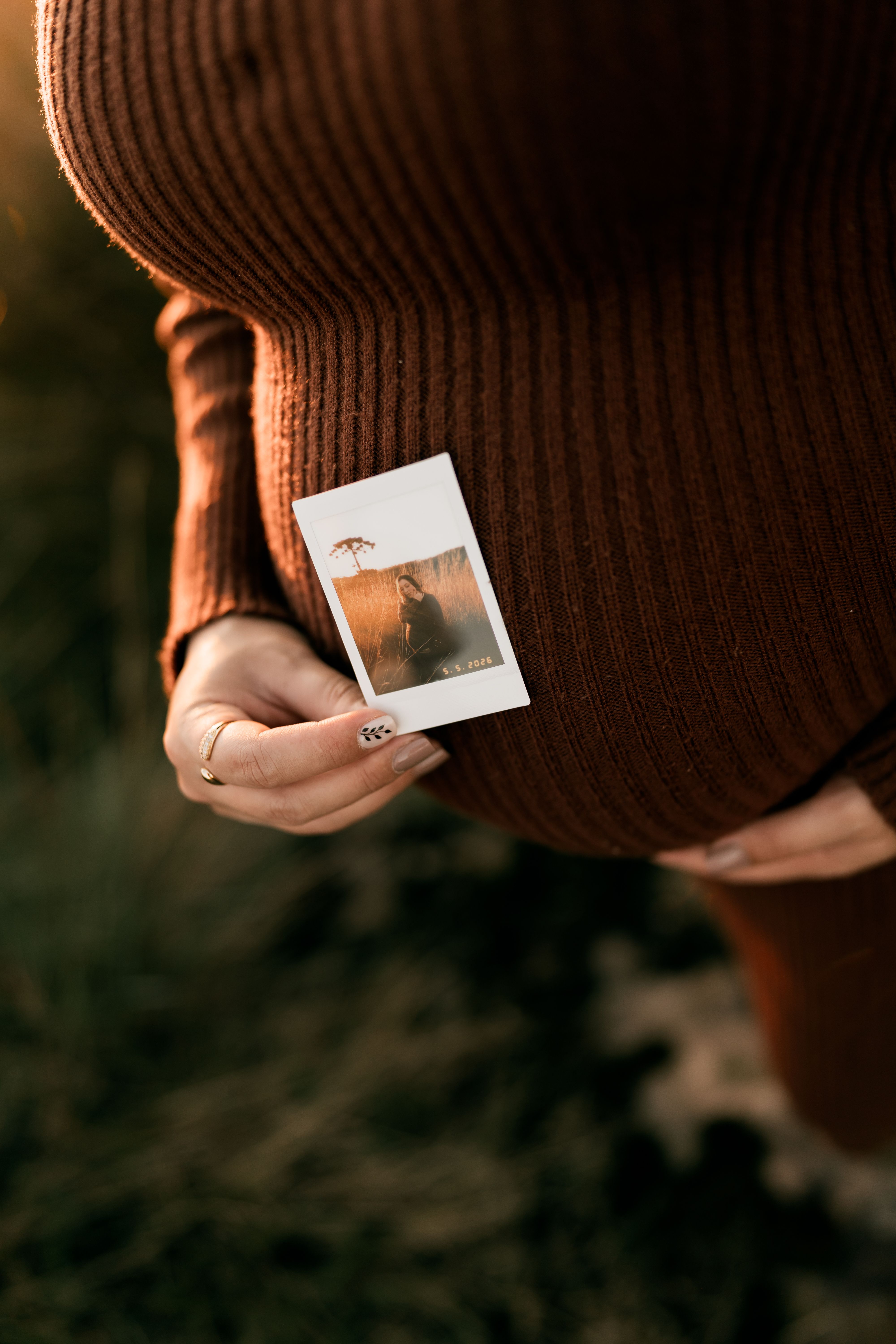 Close-up of a pregnant woman holding a small Polaroid in a serene outdoor setting.