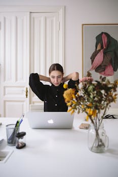 A woman focuses on work at a laptop in a minimalist home office setting with a flower vase.