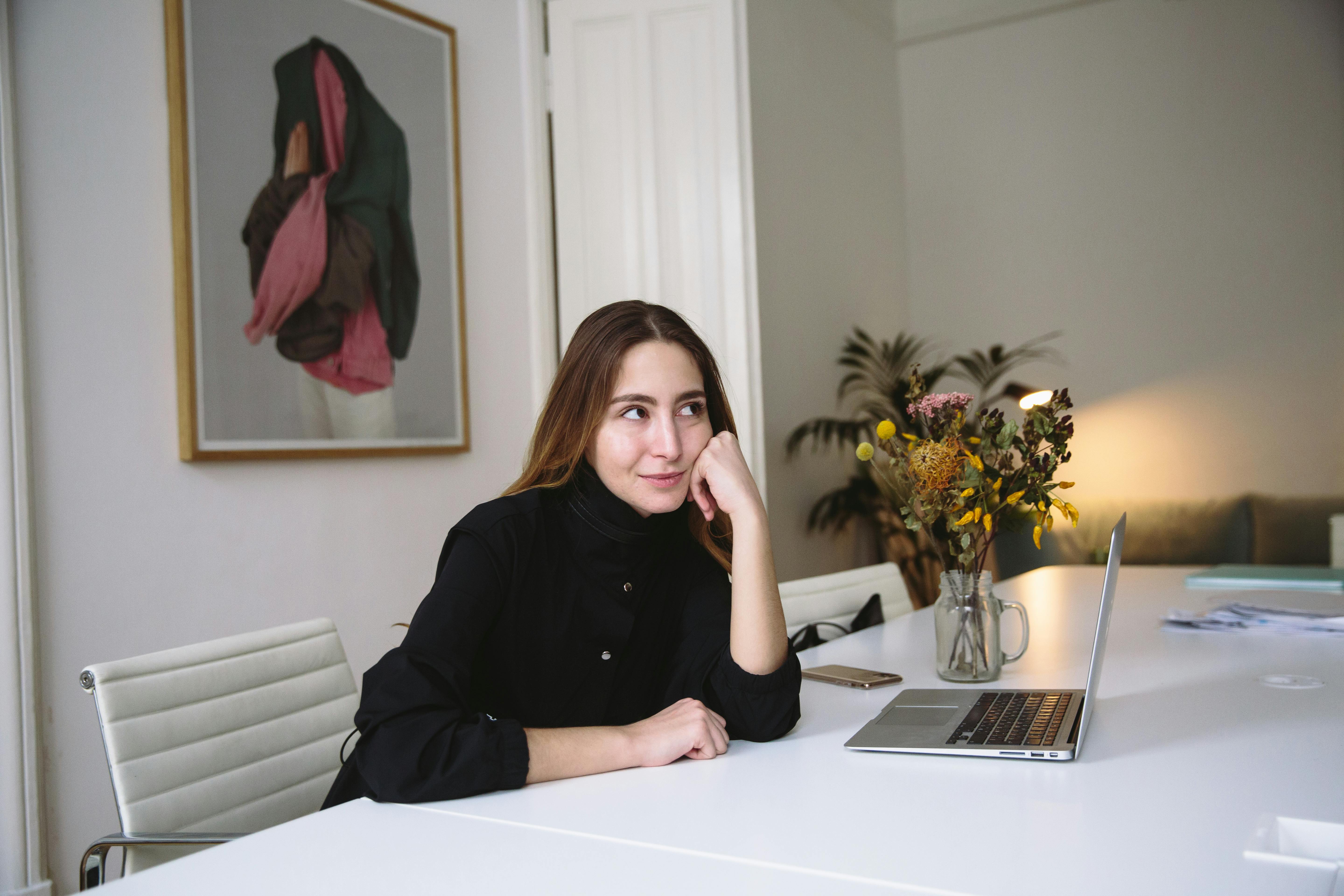 一位年轻女子坐在现代办公室的办公桌前，手里拿着笔记本电脑和鲜花，陷入了深思