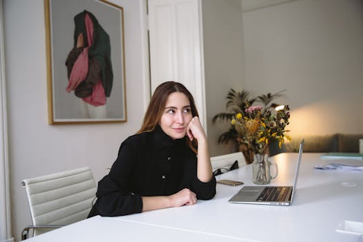 一位年轻女子坐在现代办公室的办公桌前，手里拿着笔记本电脑和鲜花，陷入了深思