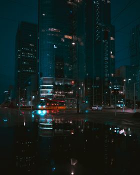 Dramatic cityscape at night featuring high-rise buildings and vibrant street reflections.
