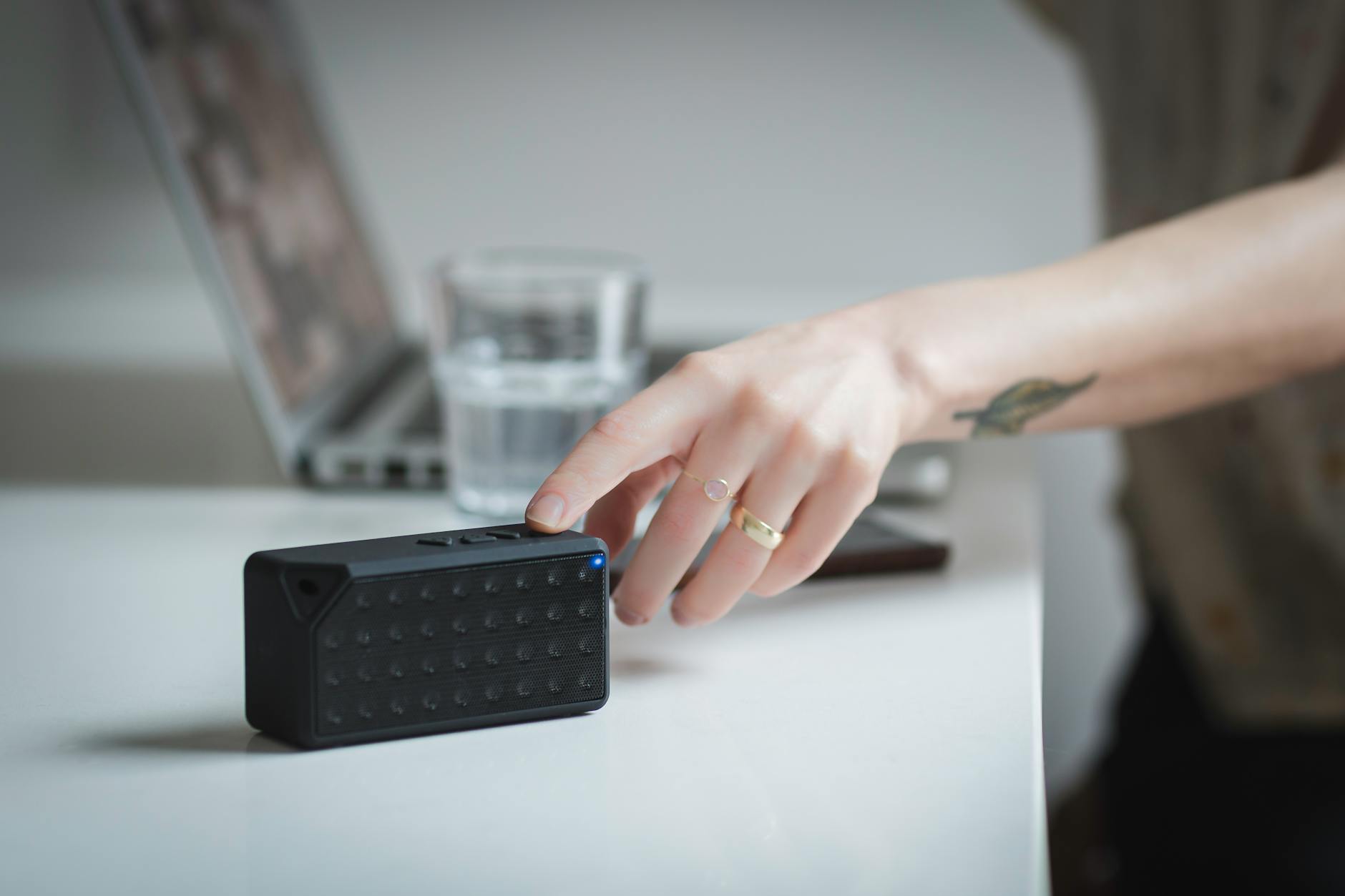Close-up of a person adjusting a portable Bluetooth speaker on a desk indoors, with a laptop in the background.