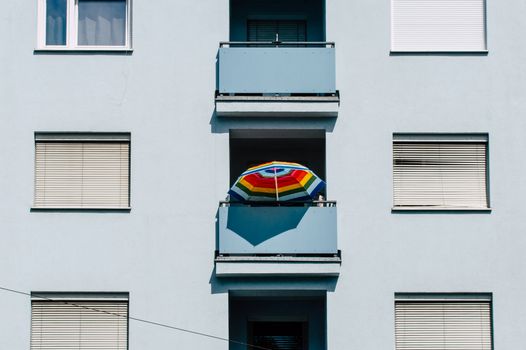 Vibrant rainbow umbrella stands out on a blue urban apartment building balcony.