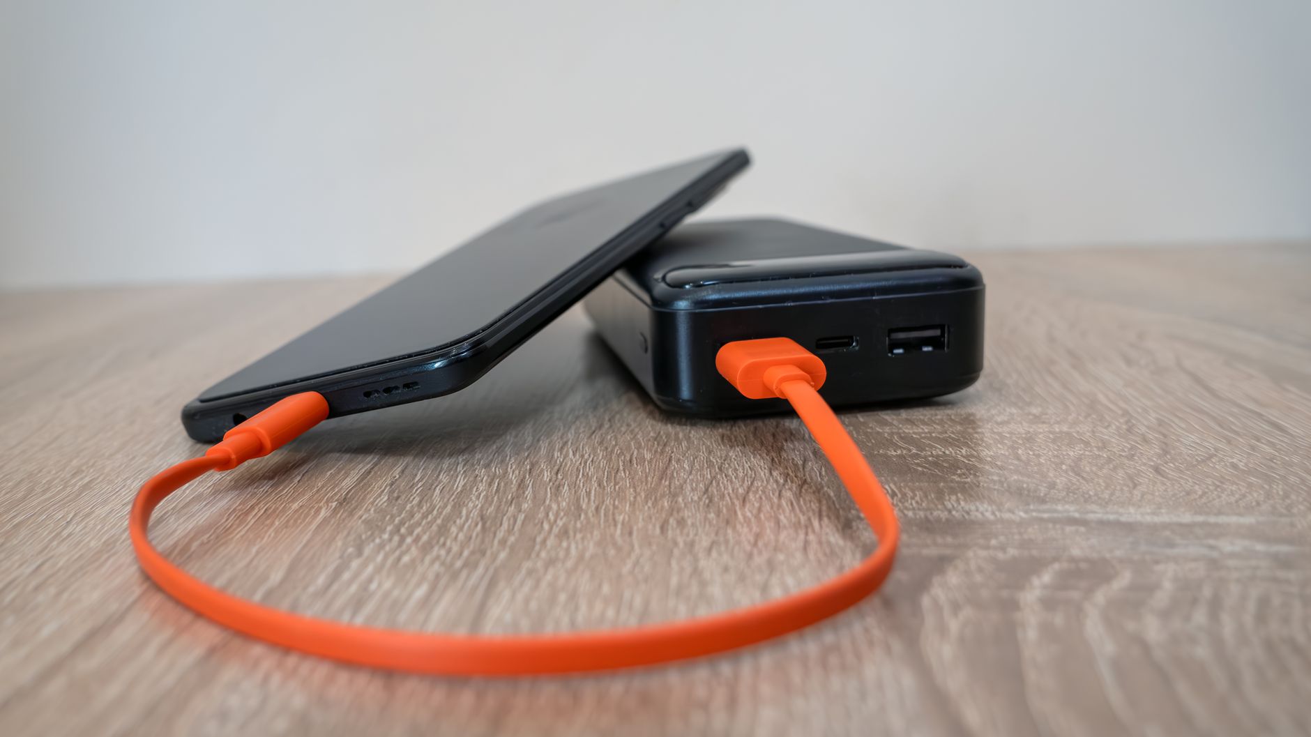 A smartphone charging via an orange cable connected to a power bank on a wooden surface.
