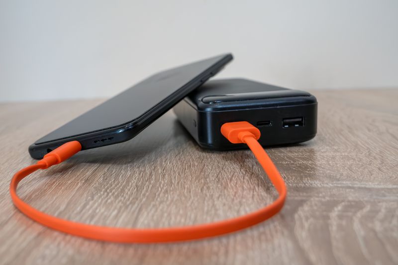 A smartphone charging via an orange cable connected to a power bank on a wooden surface.