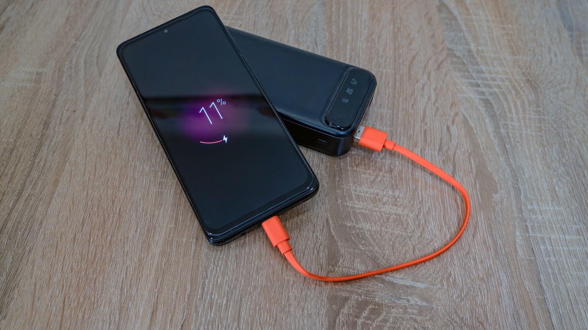 A smartphone charging from a power bank with an orange cable on a wooden table.
