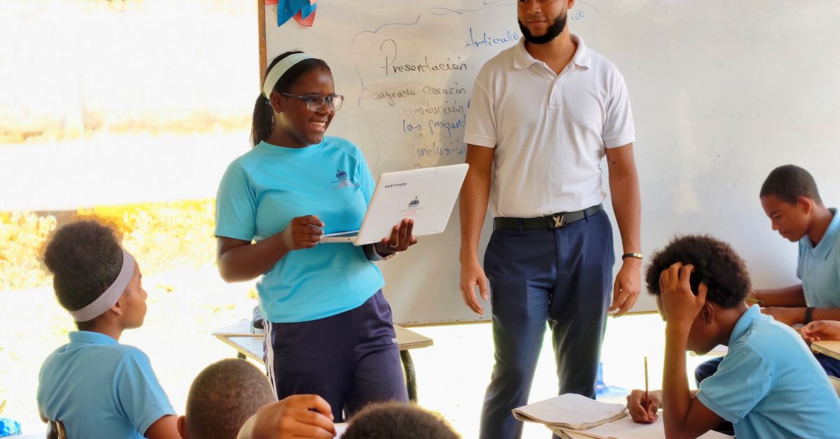 Teacher standing at a whiteboard with a laptop open to ChatGPT, smiling, classroom in background