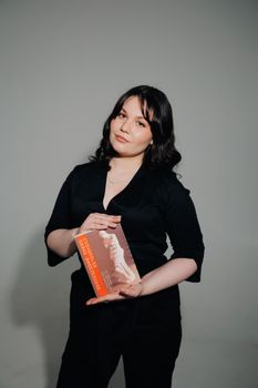 Young woman in black outfit holding a book, posing confidently in a studio setting.