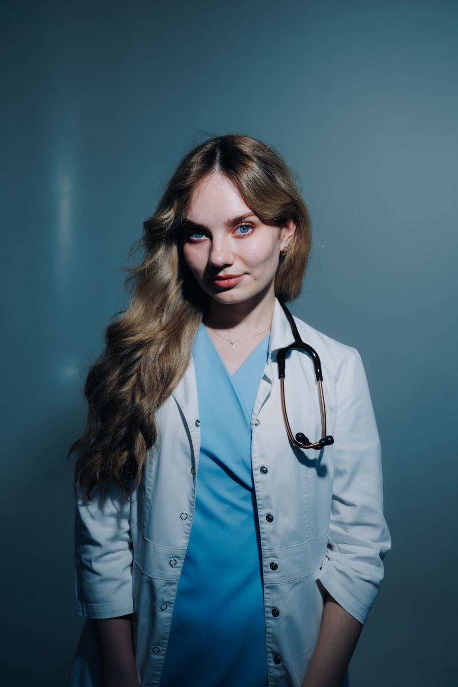 Clinician in a white coat and stethoscope, lit against a deep blue backdrop — photo by Daniil Kondrashin on Pexels