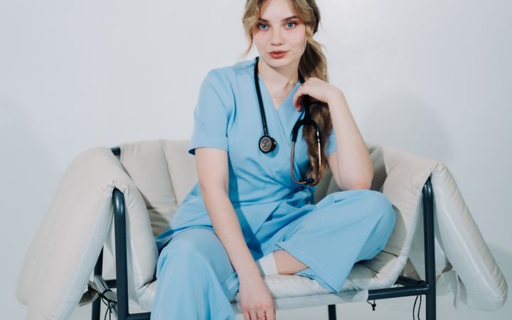Portrait of a young female nurse in blue scrubs sitting on a chair with a stethoscope.