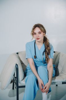 A young nurse in blue scrubs and stethoscope sitting on a chair indoors.