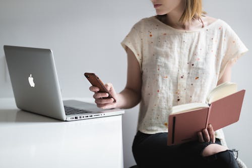 Woman Holding Book and Iphone Beside Macbook Pro
