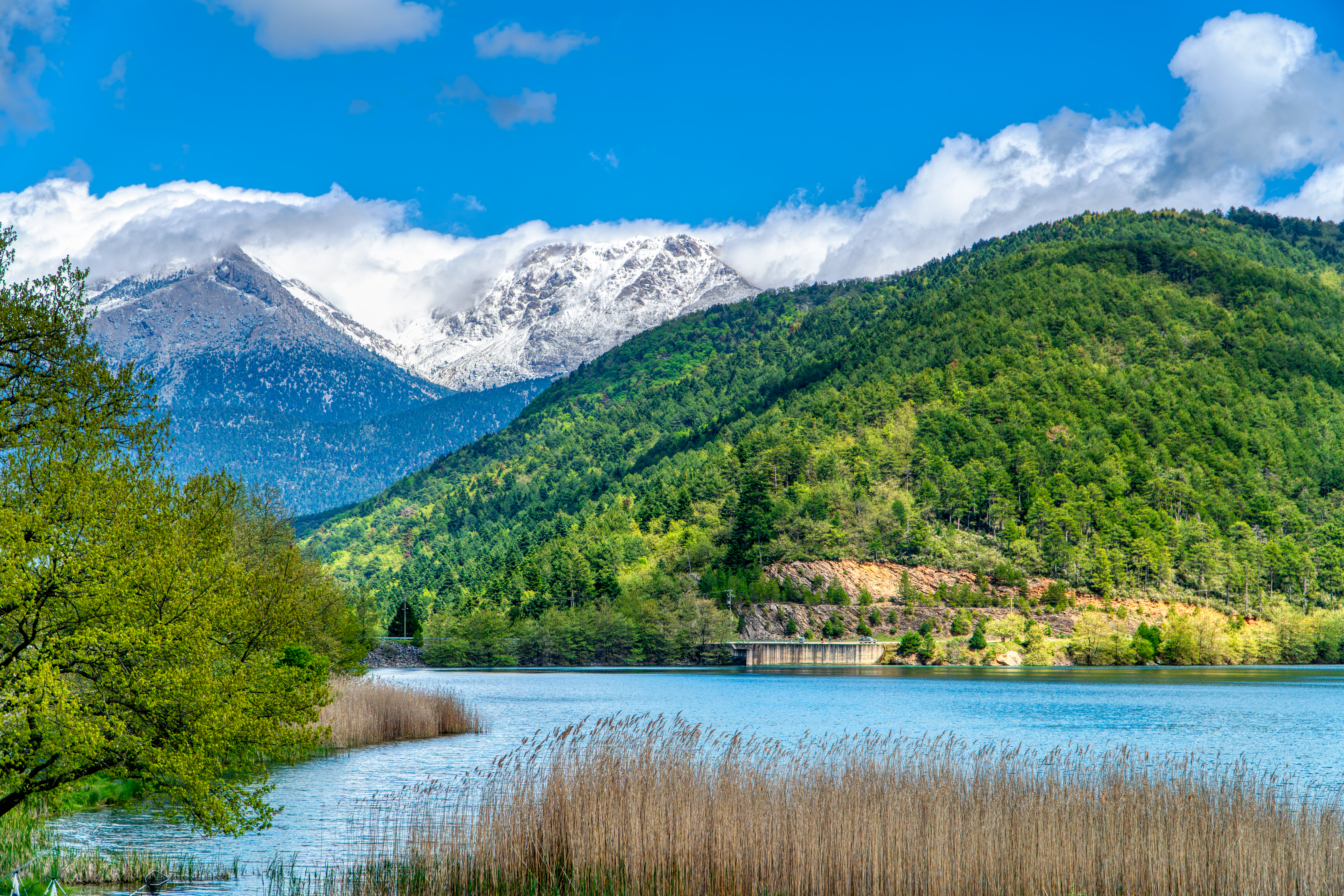 Experience the serene beauty of Lake Doxa surrounded by lush greenery and snow-capped mountains in Greece.