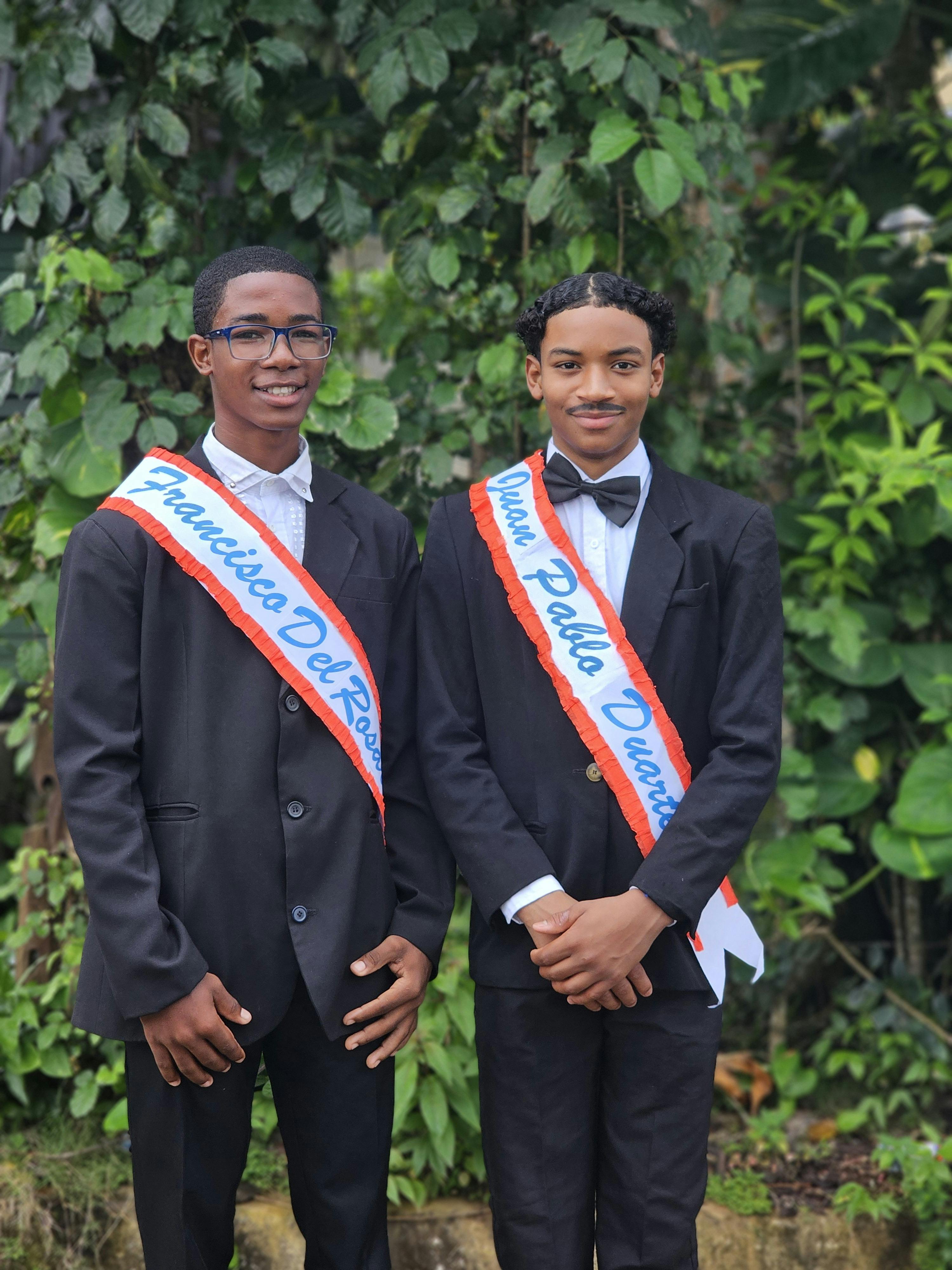Two young men posing outdoors in suits with sashes. They stand smiling against a lush green background.